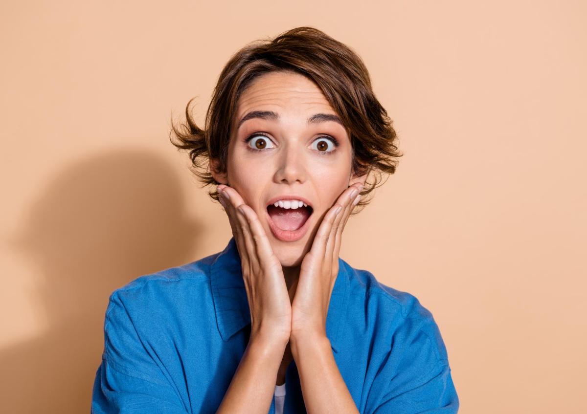 A woman with short hair holds her face with both hands, eyes wide open and mouth open, expressing surprise or amazement.