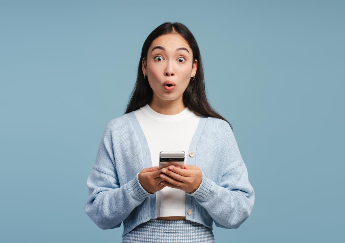 Vrouw kijkt verbaasd met grote ogen en ronde mond, houdt een telefoon vast met beide handen.