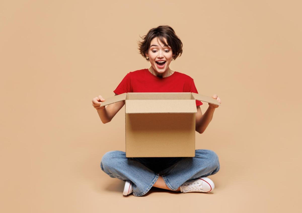 A young woman sits cross-legged on the floor, holding an open cardboard box in front of her with an expression of surprise and excitement.
