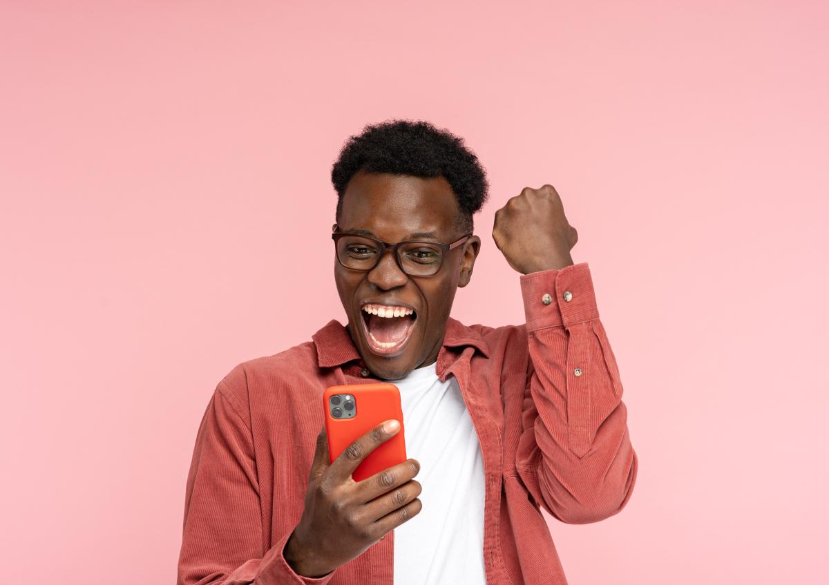 A joyful man wearing glasses looks at his phone with excitement, raising his fist in celebration.