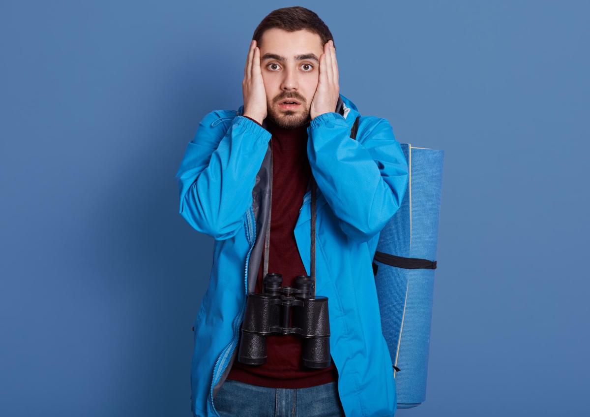 A man wearing outdoor gear looks shocked or worried, holding his face with both hands. He has binoculars hanging around his neck and a rolled-up mat strapped to his back.