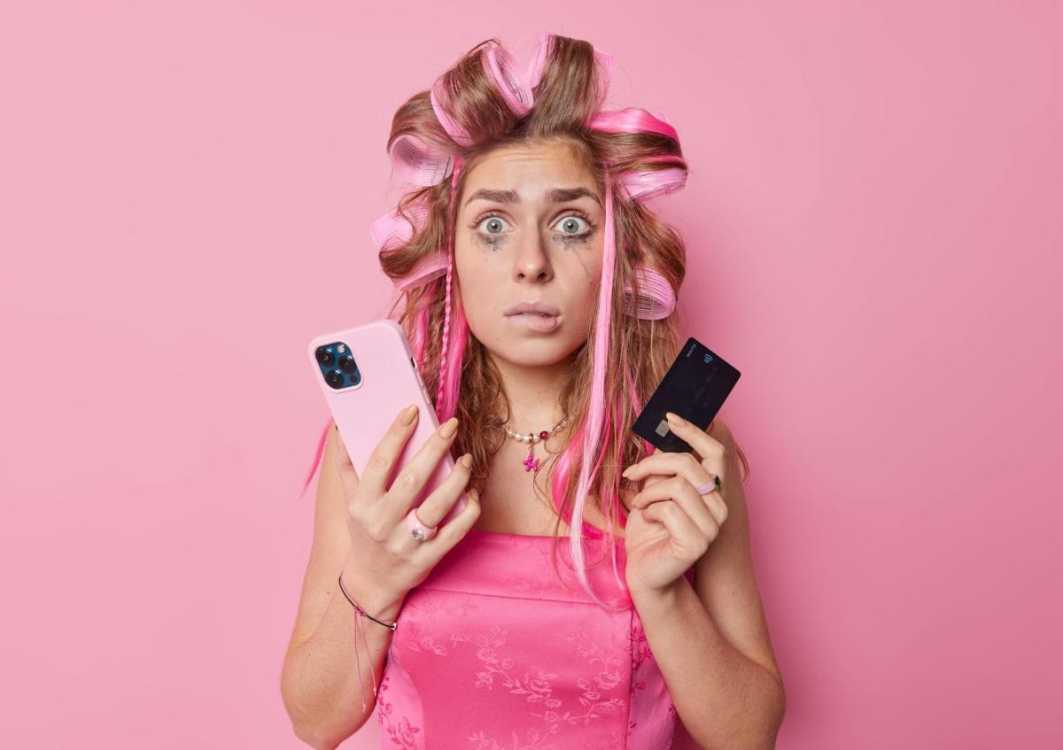 A young woman with hair curlers and smudged makeup holds a smartphone in one hand and a credit card in the other, looking confused and distressed.