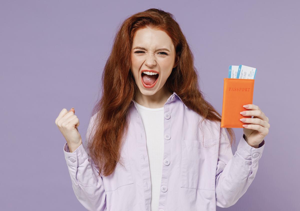 A young woman with long red hair shows excitement and triumph, holding a passport holder with flight tickets visible while making a fist with her other hand.