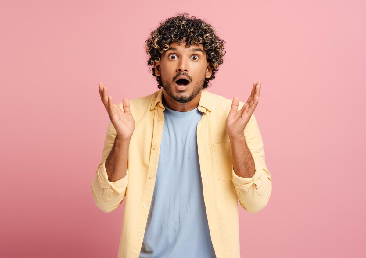 Een man met krullend haar steekt beide handen op met de handpalmen naar buiten. Zijn ogen zijn wijd open en zijn mond staat een beetje open, wat verrassing of schok uitdrukt.