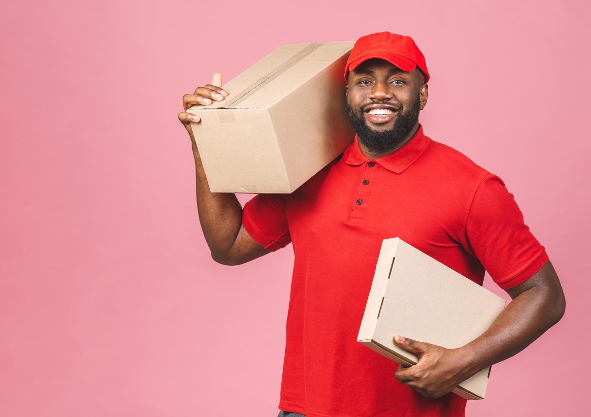 A smiling delivery man wearing a cap holds two cardboard boxes, one on his shoulder and the other in his hand, conveying readiness and friendliness.