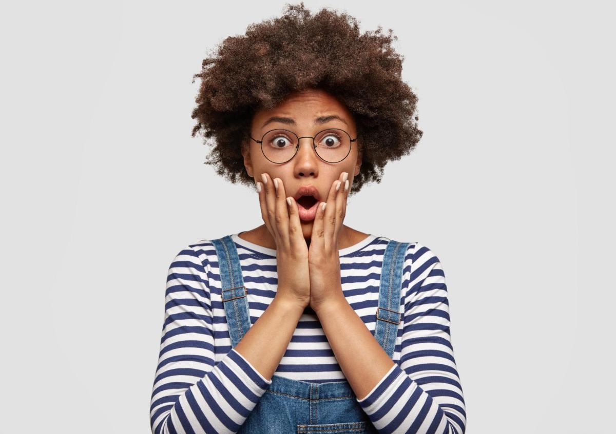 Een vrouw met een bril houdt haar gezicht vast met beide handen. Haar mond is open en ze kijkt verrast of geschrokken.