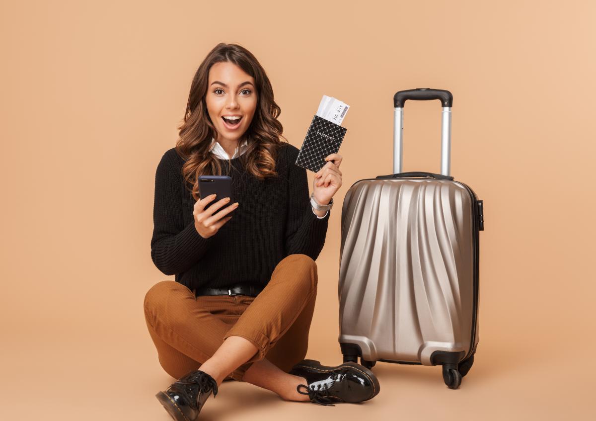 A young woman sitting cross-legged on the floor next to a suitcase, looking excited and surprised, holding a phone in one hand and a passport with a boarding pass in the other.