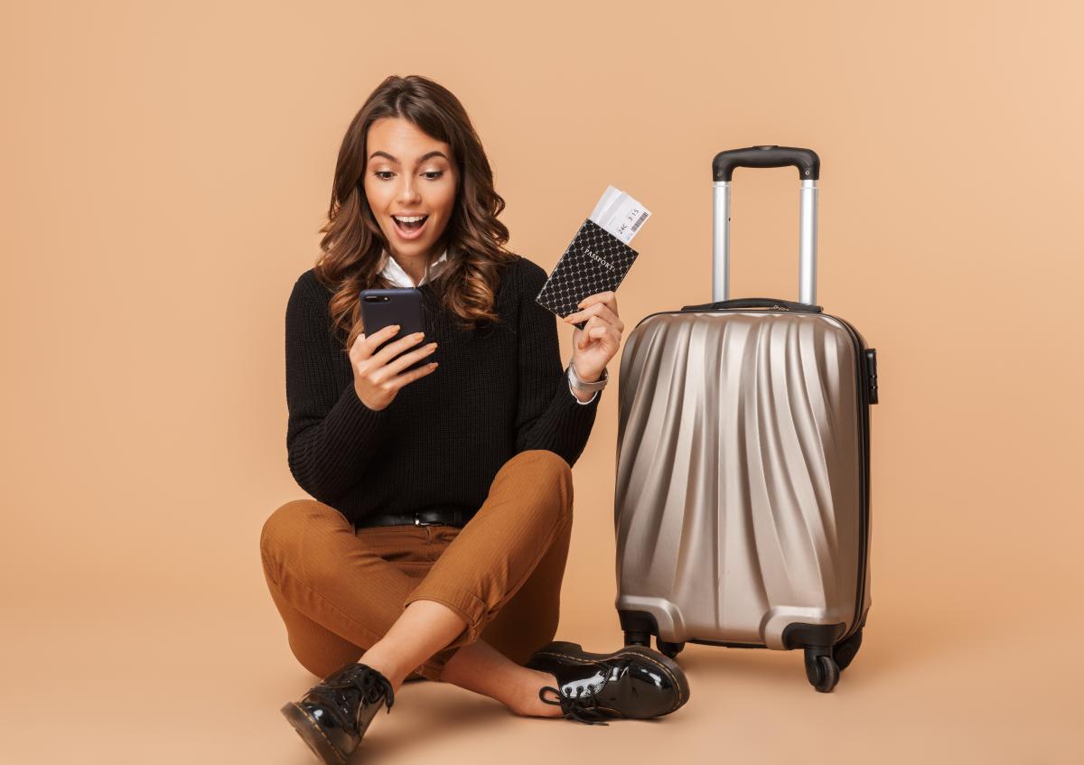 A woman sits on the floor with her legs crossed, holding a smartphone in one hand and a passport with boarding passes in the other, showing a look of surprise and excitement. Next to her is a wheeled suitcase.