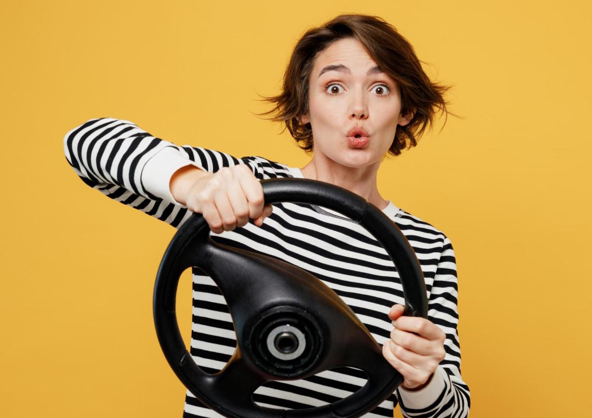A woman holding a steering wheel with both hands, making a surprised or shocked face.