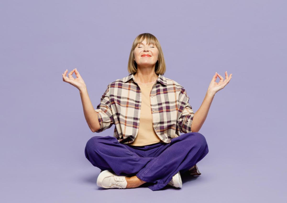 Een vrouw zit met gekruiste benen op de grond in een meditatieve houding. Haar ogen zijn gesloten en ze glimlacht rustig, terwijl ze haar handen ontspannen omhoog houdt met duim en wijsvinger tegen elkaar, alsof ze aan het mediteren is.