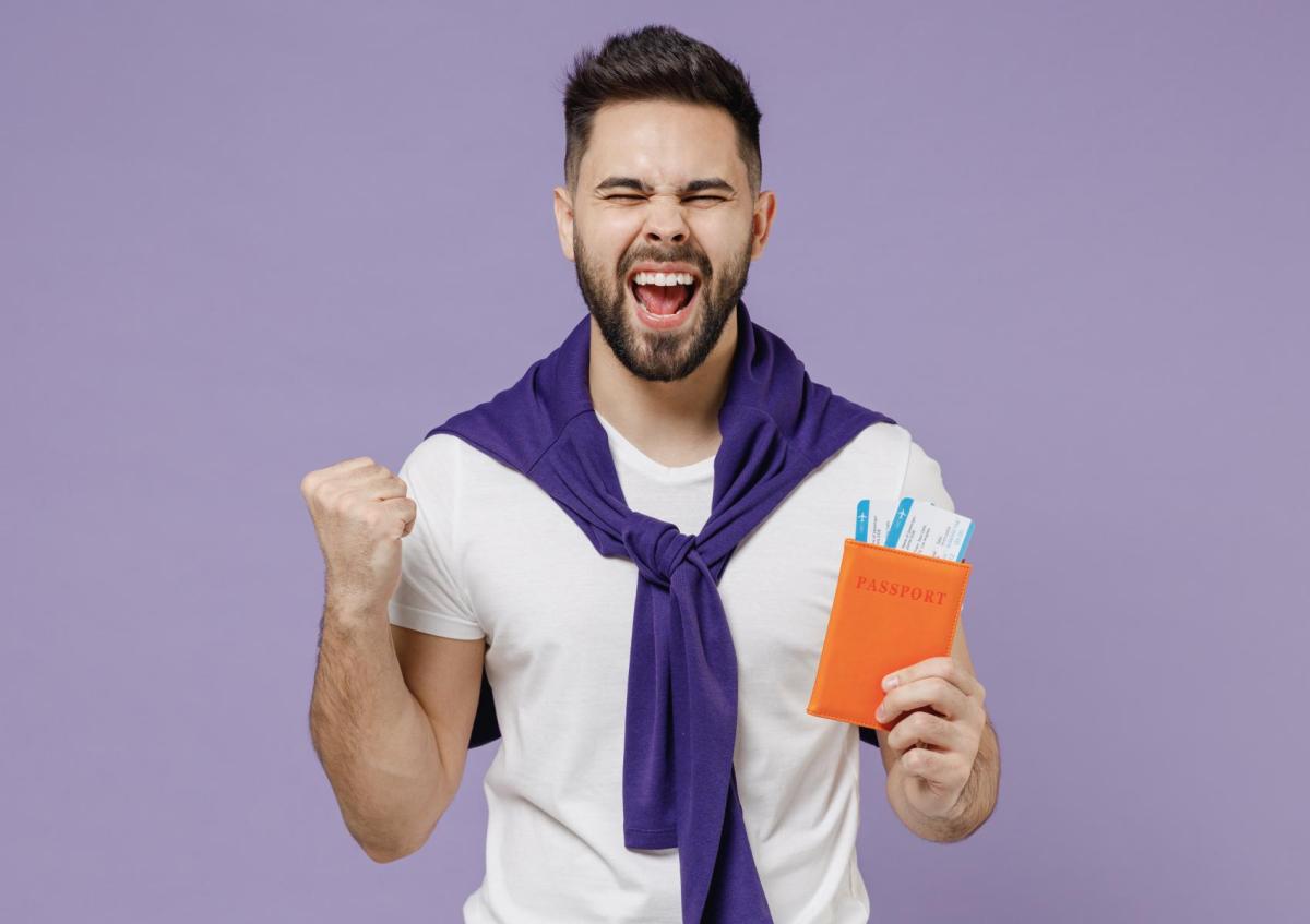 Een blije jonge man met een baard houdt een paspoorthoes vast met daarin twee instapkaarten. Met zijn andere hand maakt hij een feestelijk gebaar en zijn mond is open van blijdschap. 