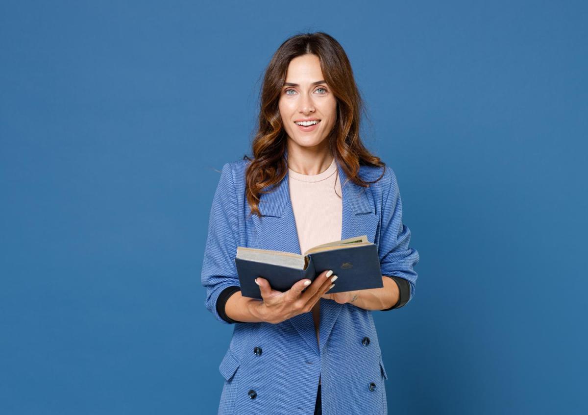 Een vrouw staat met een open boek in haar handen, kijkt met een lichte glimlach en een nieuwsgierige of bedachtzame blik naar de camera.