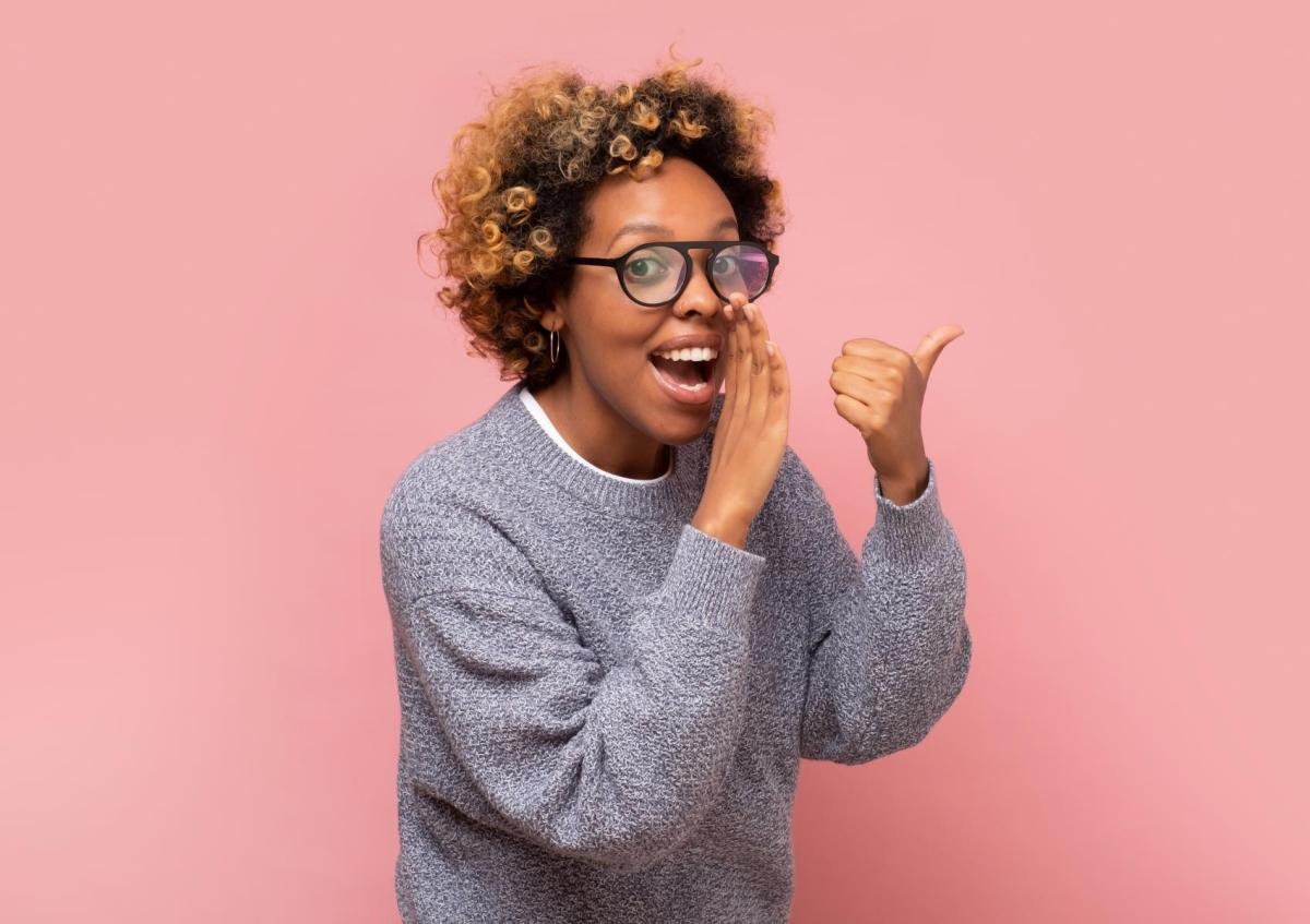 Vrouw met open mond alsof ze iets vertelt of roept, kijkt enthousiast. Ze houdt één hand voor haar mond en steekt met de andere hand haar duim omhoog.
