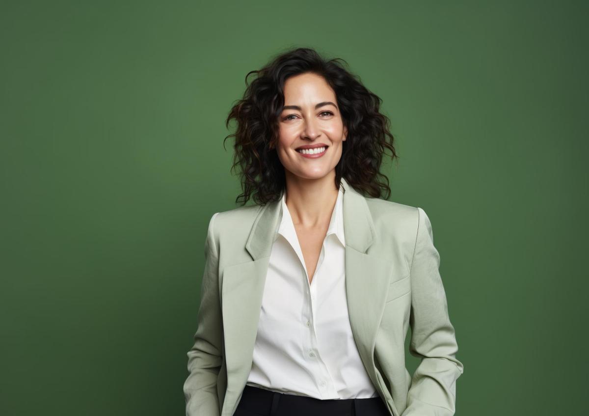 Vrouw kijkt zelfverzekerd en vriendelijk naar de camera met een brede glimlach. Ze staat rechtop en heeft haar handen ontspannen in haar zakken.