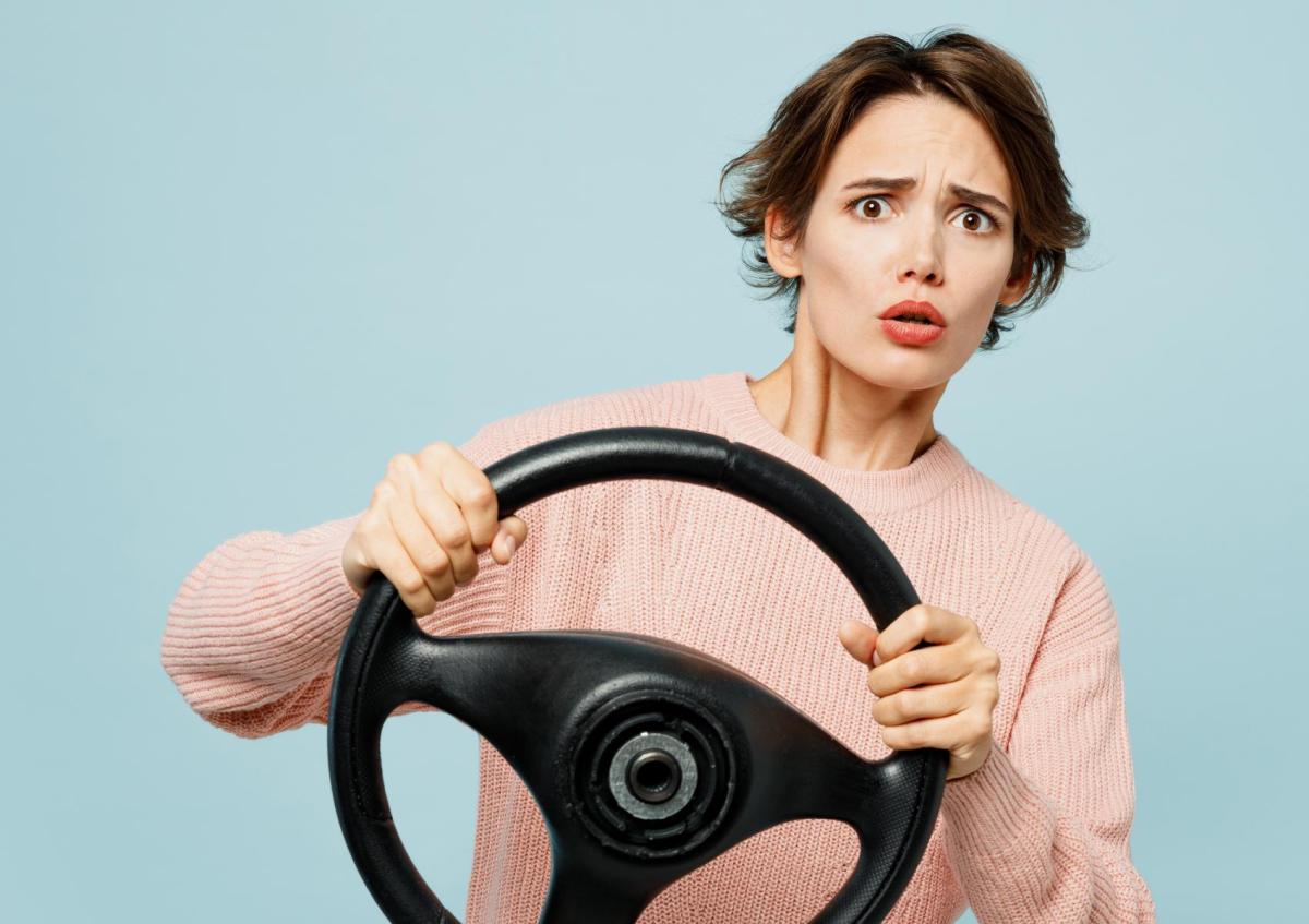 Een vrouw met een bezorgde of geschrokken blik houdt een stuur vast alsof ze aan het autorijden is. Ze kijkt recht voor zich uit met opgetrokken wenkbrauwen en een open mond.