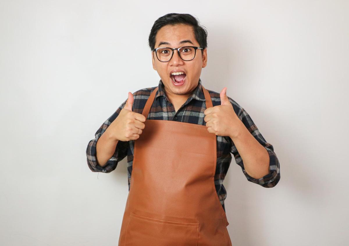 Een man met een bril en draagt een schort. Hij kijkt blij en enthousiast, met zijn mond open en geeft met beide handen een duim omhoog.