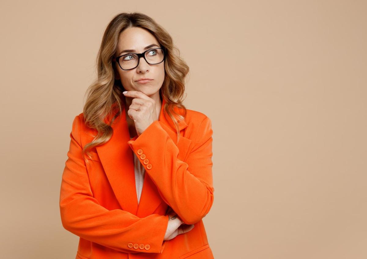 Vrouw kijkt nadenkend naar boven rechts met opgetrokken wenkbrauwen en licht gespannen mond. Ze houdt één hand aan haar kin en haar andere arm is gekruist voor haar lichaam.