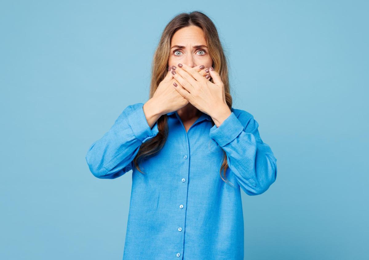 Vrouw kijkt geschrokken of bezorgd met grote ogen naar de camera. Ze bedekt haar mond met beide handen.