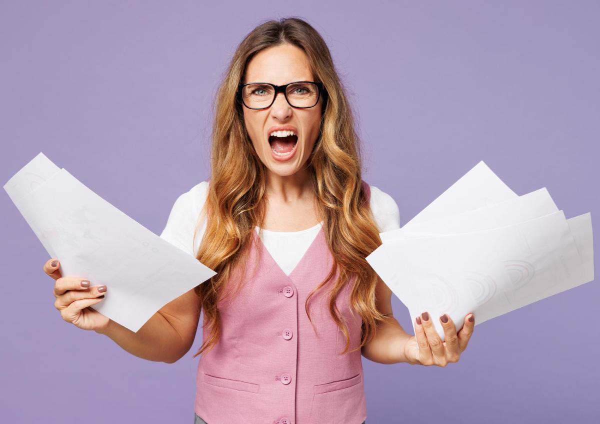 Vrouw kijkt boos en gefrustreerd met open mond. Ze houdt een stapel papieren in beide handen omhoog.