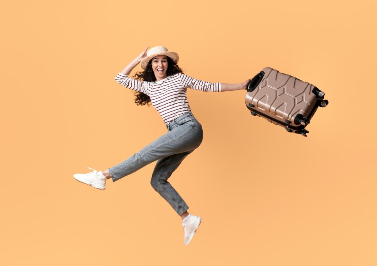 A joyful young woman holding a large tan rolling suitcase by its handle and smiling brightly