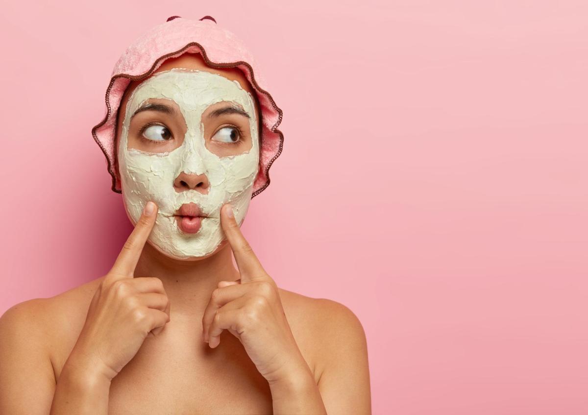 Vrouw met gezichtsmasker kijkt nieuwsgierig en speels opzij terwijl ze met haar wijsvingers haar gezicht aanraakt.