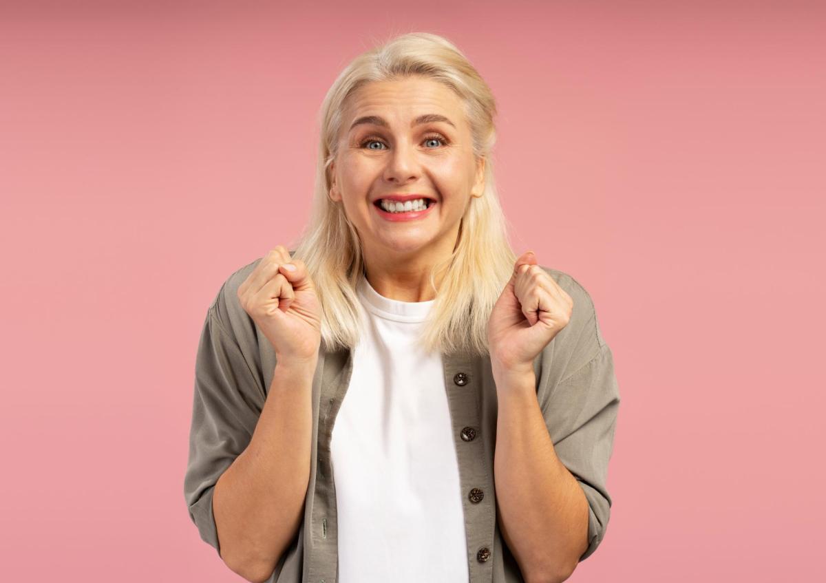 Vrouw kijkt blij en enthousiast, met een brede glimlach en opgetrokken handen in een feestelijke of opgewonden houding.