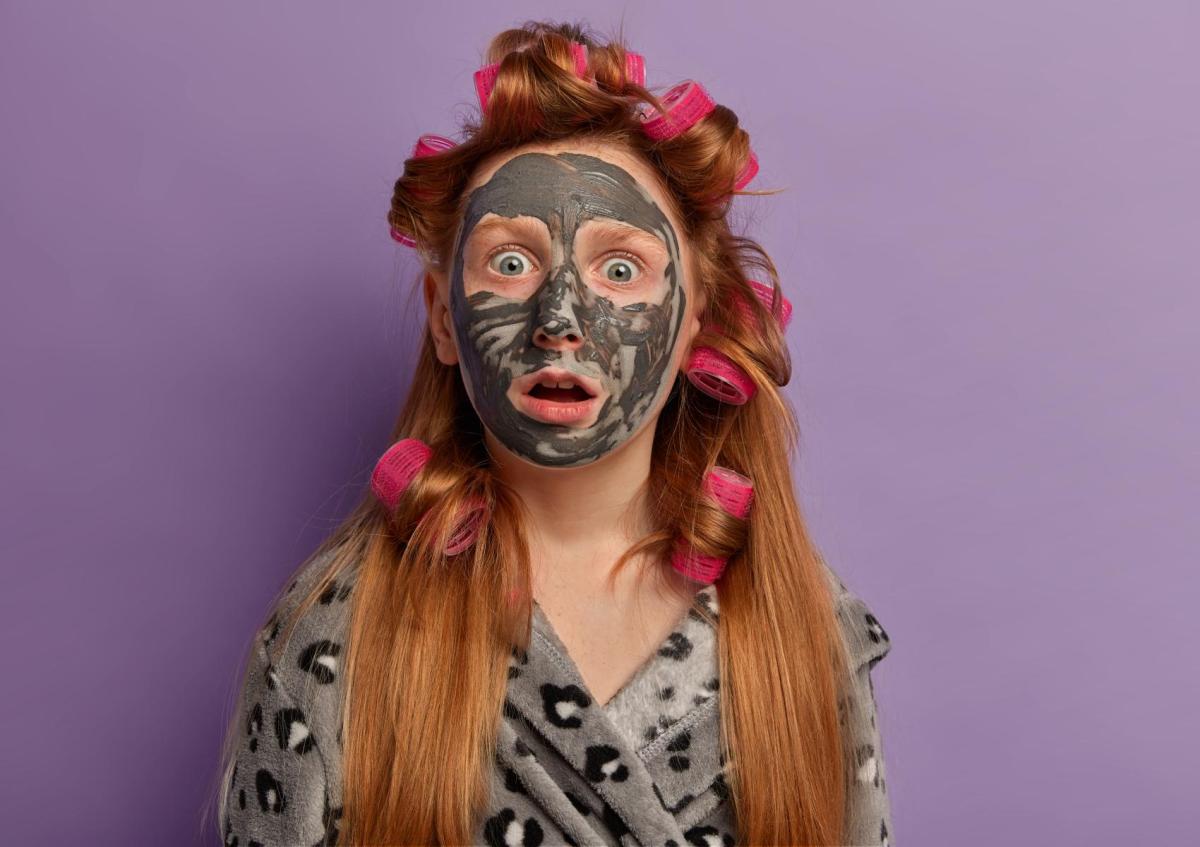 Vrouw kijkt verrast en verbaasd met haar mond iets open en ogen wijd open. Ze heeft haar hoofd bedekt met een kleimasker en draagt haar haar in krulspelden.