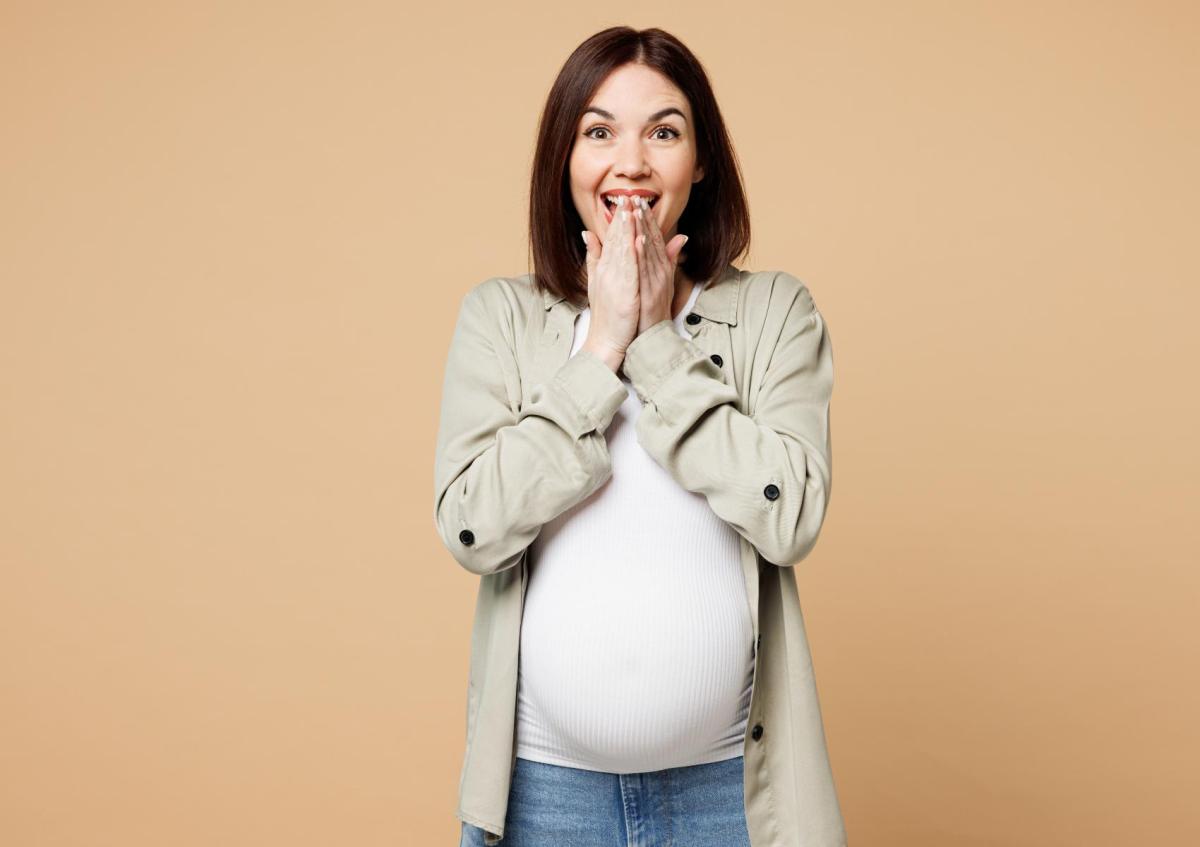 Zwangere vrouw kijkt blij en verrast, ze houdt haar handen voor haar mond en haar ogen zijn wijd open. Ze lijkt net een onverwacht leuk nieuws te hebben gehoord.