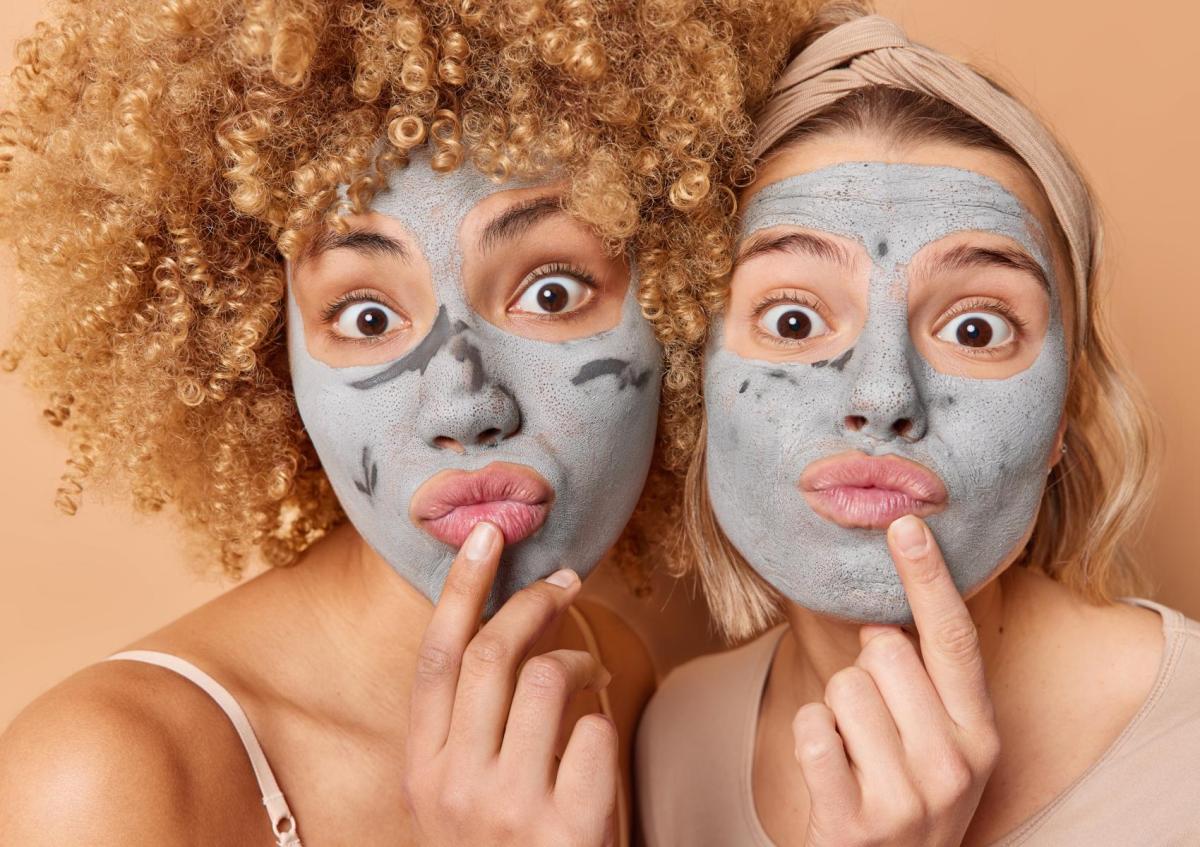 Twee vrouwen met gezichtsmaskers op, trekken een speelse, overdreven verbaasde gezichtsuitdrukking met grote ogen en licht vooruitgestoken lippen. Beiden raken met een vinger hun kin aan, alsof ze nadenken of iets grappigs overwegen.