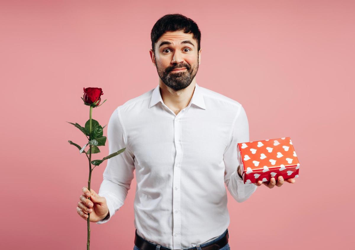 Een man met een lichte glimlach en opgetrokken wenkbrauwen kijkt direct naar de camera met een iets vragende of onzekere blik. Hij houdt in zijn rechterhand een enkele roos vast en in zijn linkerhand een cadeauverpakking met hartjesmotief, alsof hij iets aanbiedt of een keuze maakt.