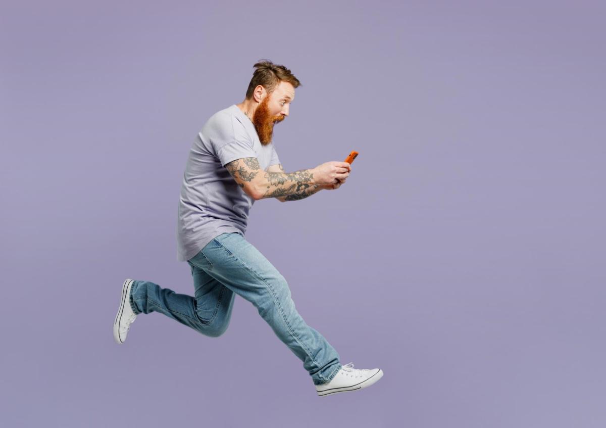 Een man die in de lucht lijkt te springen of te rennen, houdt zijn telefoon met beide handen vast en kijkt er met een grote open mond en verbaasde ogen naar, alsof hij iets onverwachts of spannends op het scherm ziet