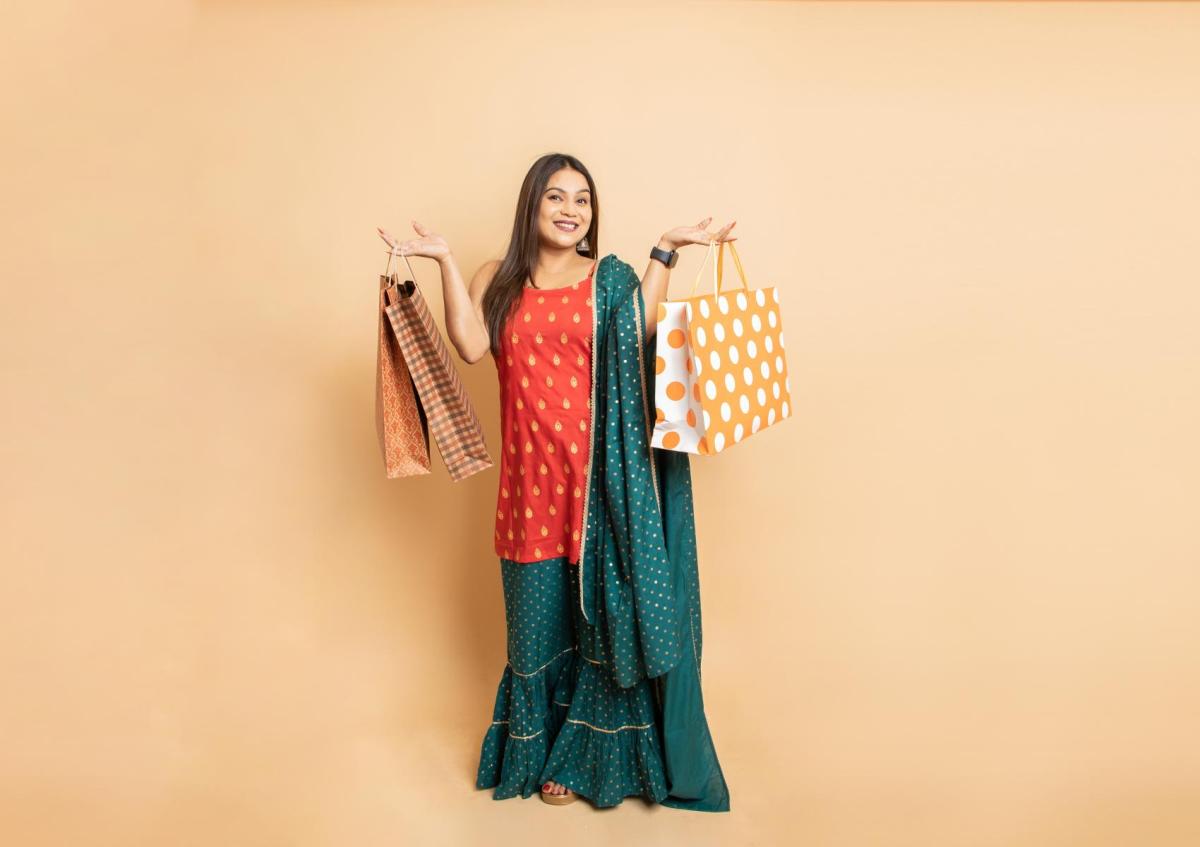 Een vrouw met een brede glimlach kijkt recht in de camera en houdt in elke hand meerdere tassen omhoog, alsof ze trots haar aankopen toont. Haar lichaamstaal straalt blijheid en tevredenheid uit.