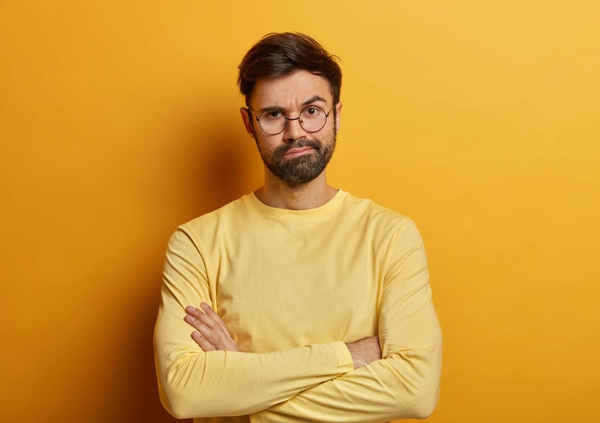 A man with beard, stands with his arms crossed. He has a slightly raised eyebrow and a skeptical or doubtful expression