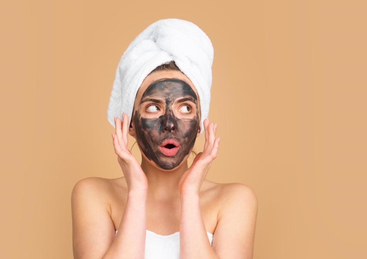 Een vrouw met een gezichtsmasker kijkt verbaasd, met grote ogen en een open mond. Ze houdt haar handen vlak bij haar gezicht, alsof ze geschrokken of verrast is.