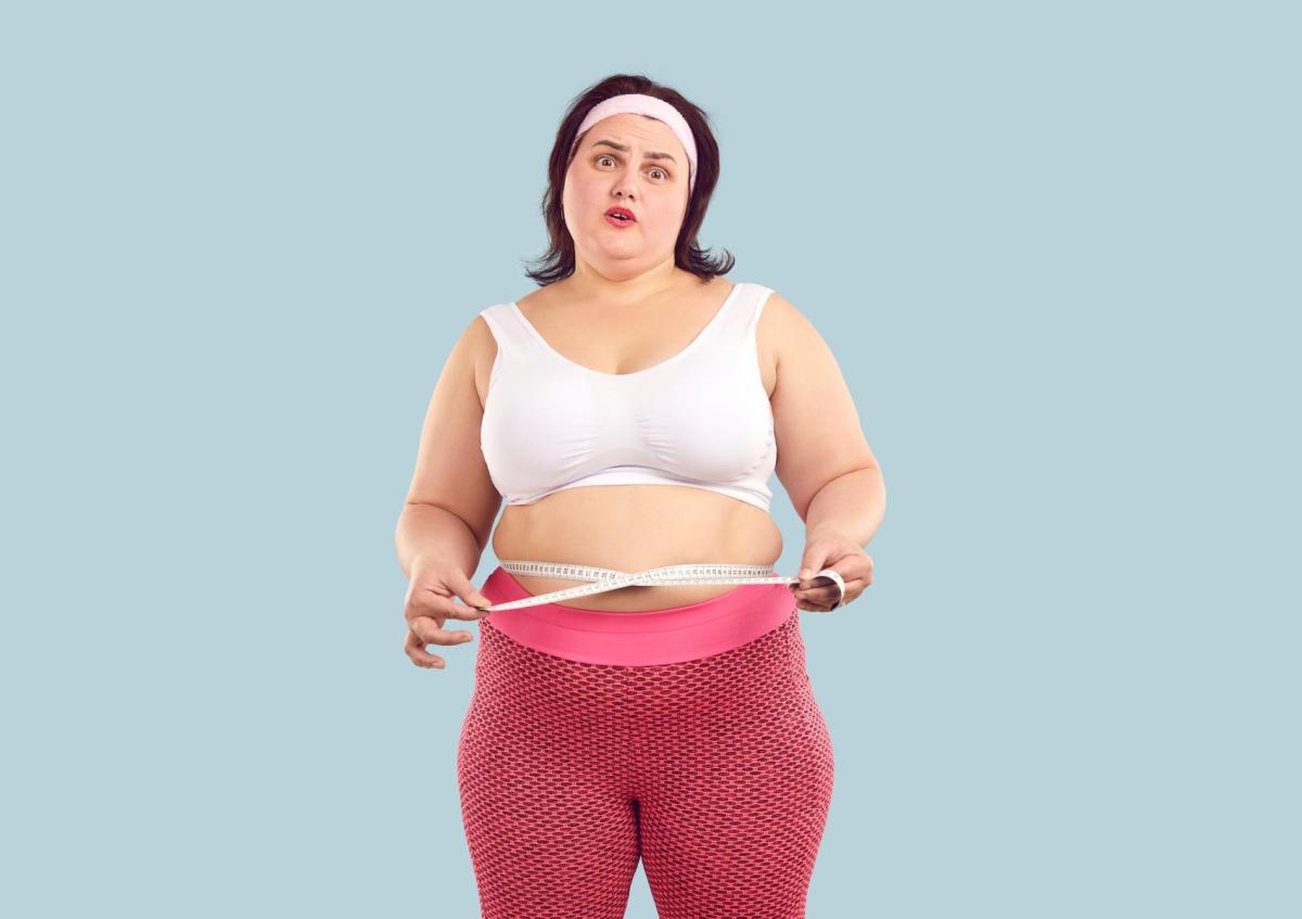 Vrouw kijkt verward en enigszins bezorgd terwijl ze met een meetlint haar taille meet. Haar mond is licht geopend en haar wenkbrauwen zijn opgetrokken, wat haar twijfelende en onzekere uitdrukking benadrukt.