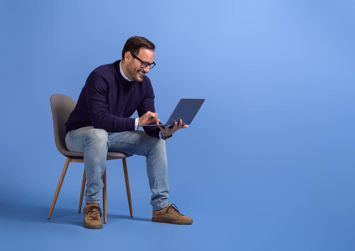 Man zit op een stoel, kijkt lachend naar zijn laptop en typt erop. Zijn gezichtsuitdrukking is vrolijk en geconcentreerd.