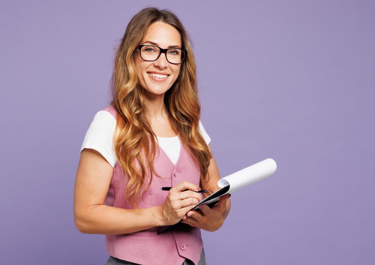 Een vrouw met een vriendelijke glimlach kijkt naar de camera terwijl ze een notitieblok vasthoudt en met een pen iets opschrijft. Ze draagt een bril en heeft lang golvend haar.