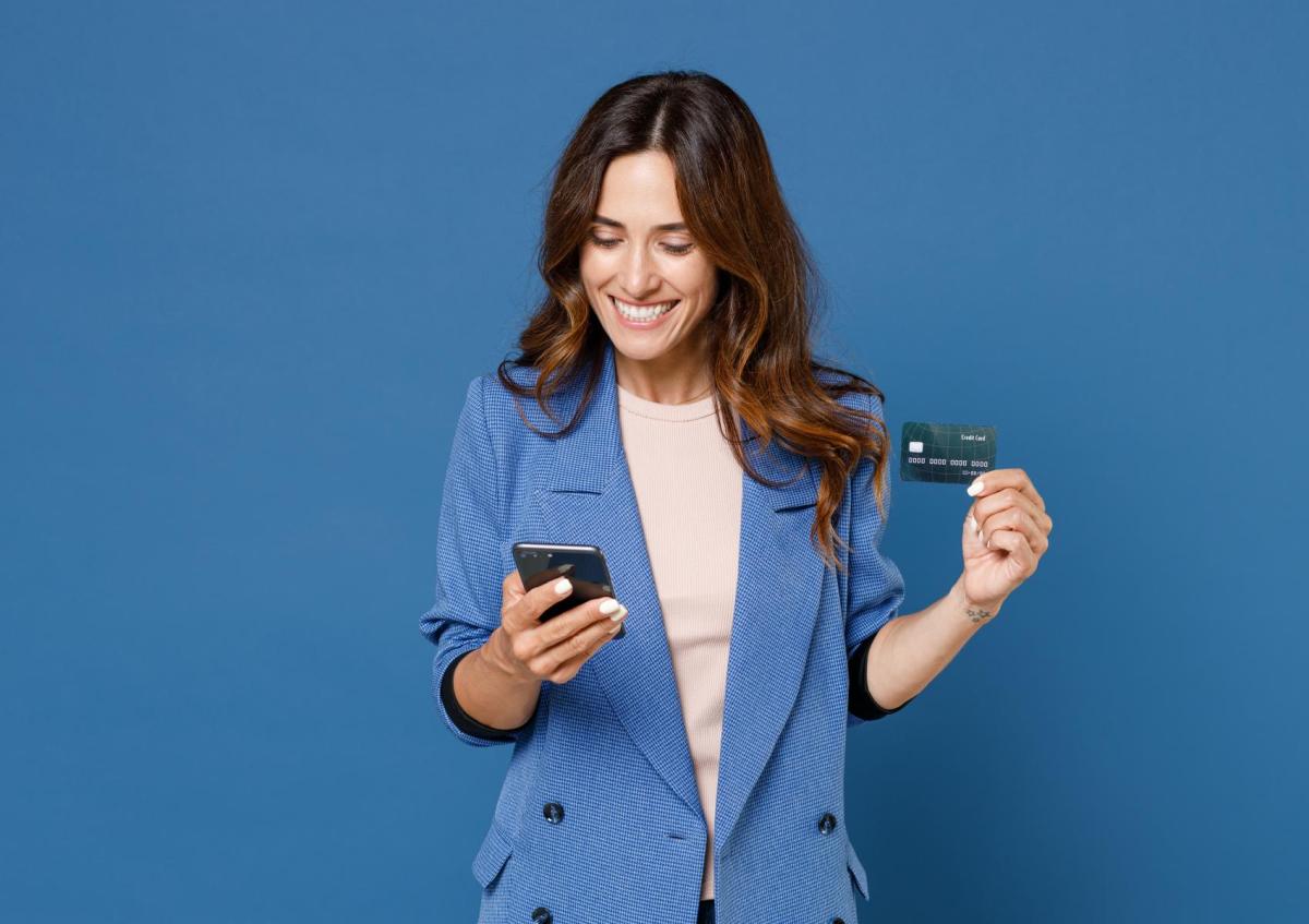 Een vrouw die glimlacht kijkt naar een mobiel apparaat in haar ene hand terwijl ze met haar andere hand een creditcard vasthoudt. Ze lijkt blij en tevreden.