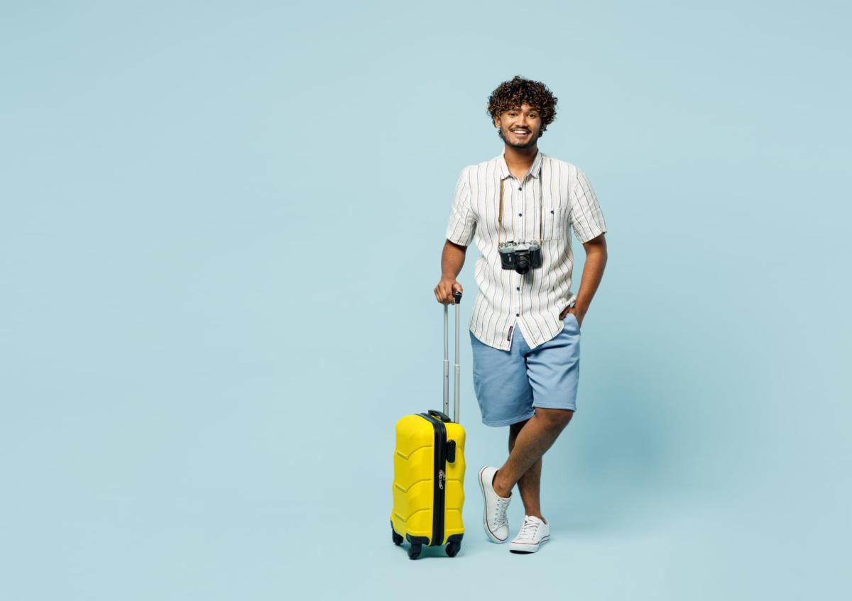 A young man standing with one leg crossed over the other, holding the handle of a wheeled suitcase with one hand and resting the other hand in his pocket. He has a camera hanging around his neck and is smiling warmly.