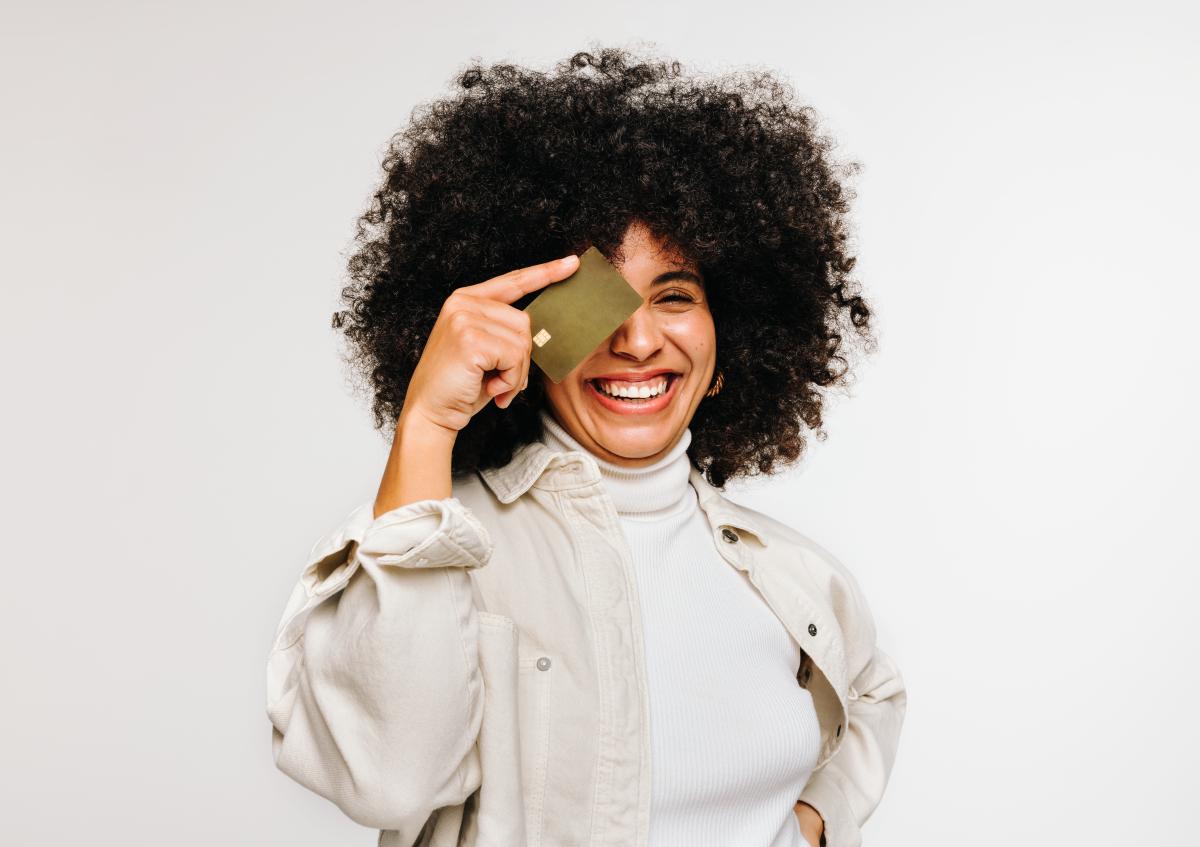 A joyful woman with curly hair smiles widely, holding a credit card up to one eye playfully.