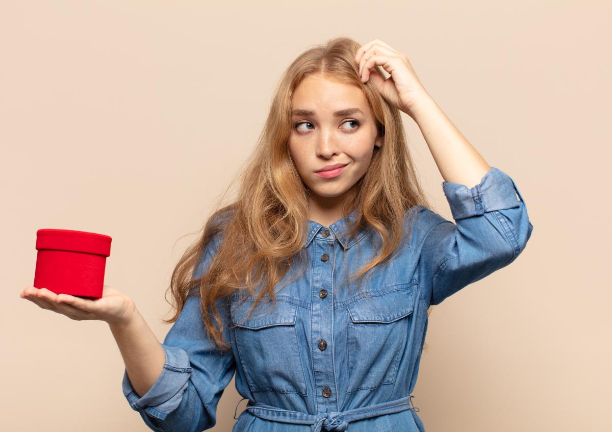 Een vrouw met een twijfelende en licht verwarde gezichtsuitdrukking krabt aan haar hoofd terwijl ze een ronde doos in haar andere hand houdt, alsof ze ergens over twijfelt of het niet zeker weet.