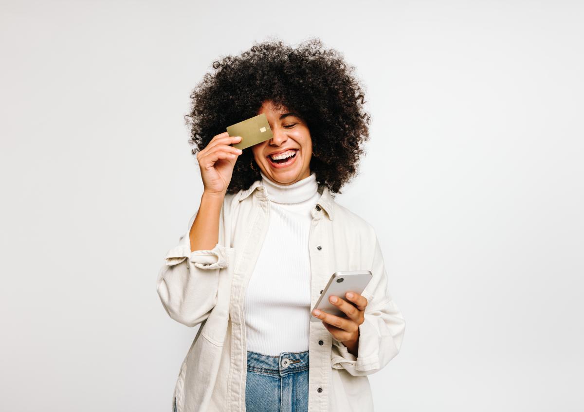 A young woman with curly hair smiles and laughs while holding a credit card up to one eye and a phone in her other hand.