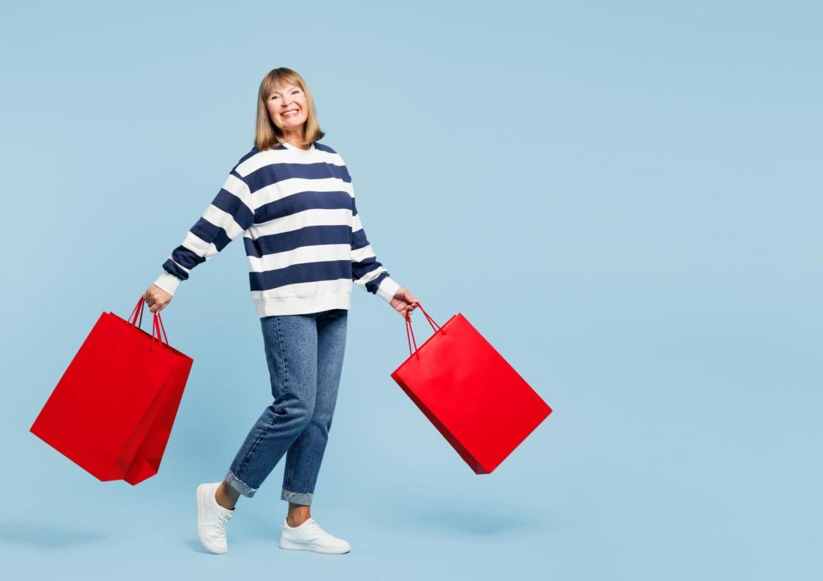 A woman, smiling and cheerful, is walking while holding two shopping bags with her arms outstretched. She appears lively and full of energy.