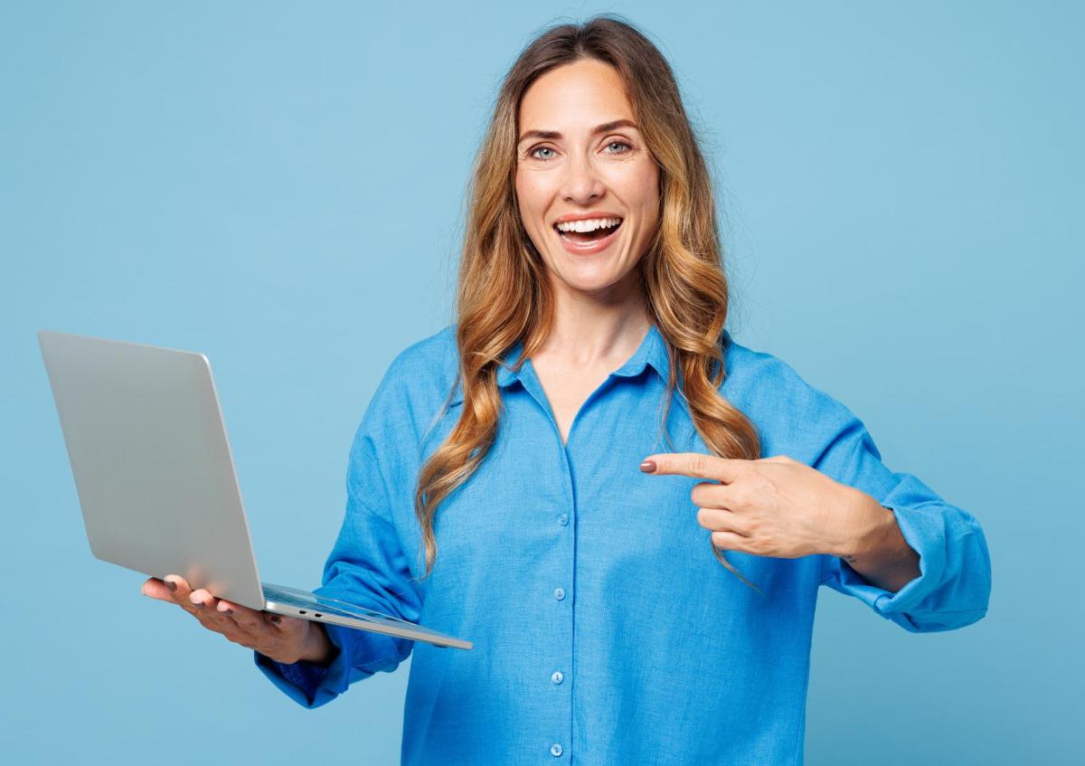 A cheerful woman with long wavy hair holds a laptop in one hand and points to it with the other, smiling confidently 
