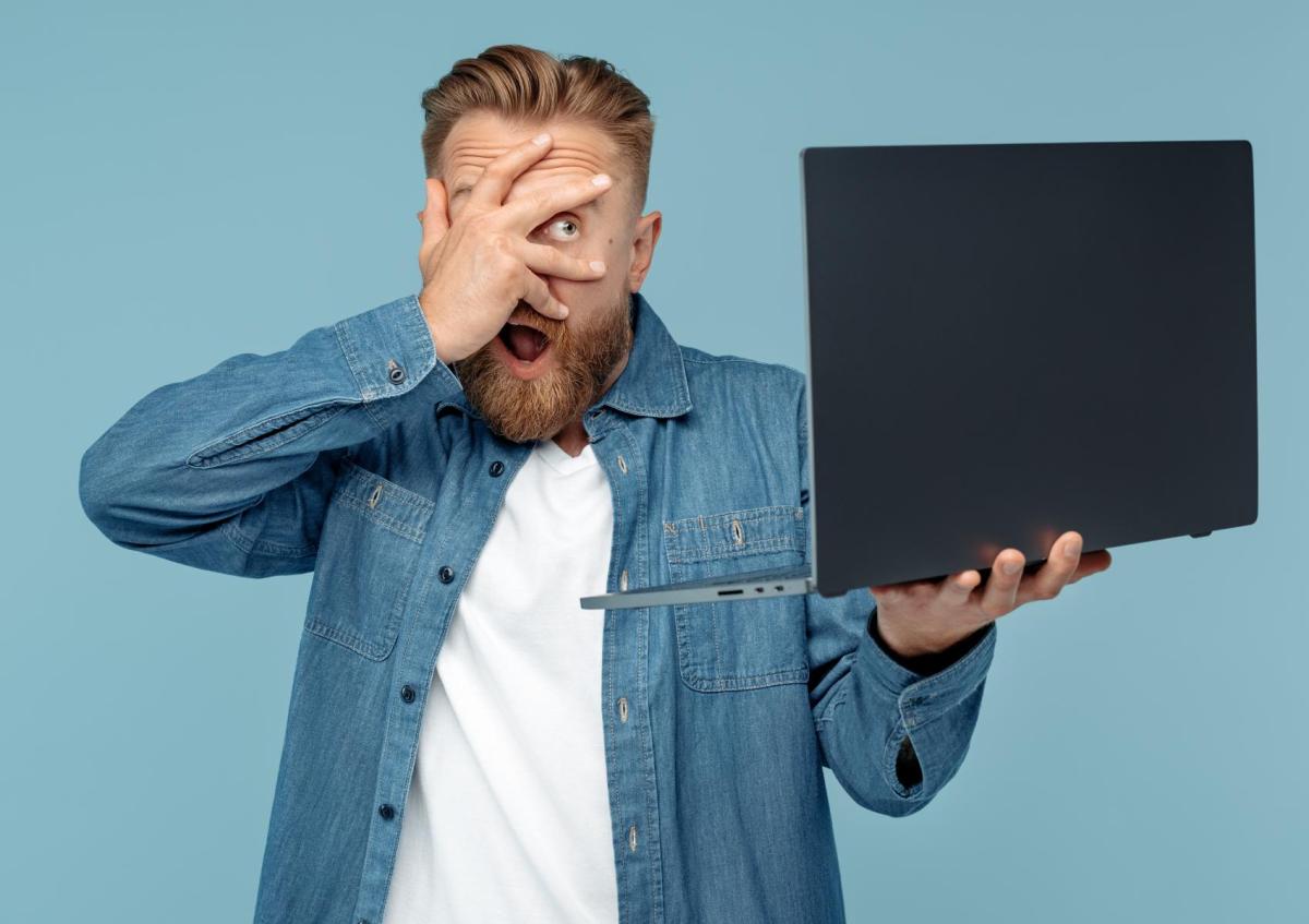 A man is holding a laptop in one hand and partially covering his face with the other, peeking through his fingers with a surprised or shocked expression.