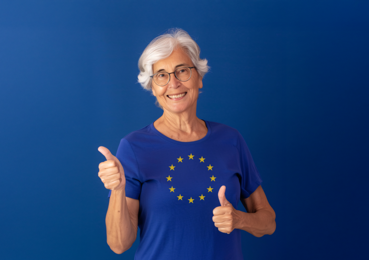 An elderly woman smiling and giving thumbs up, wearing glasses and a t-shirt featuring the European Union flag 
