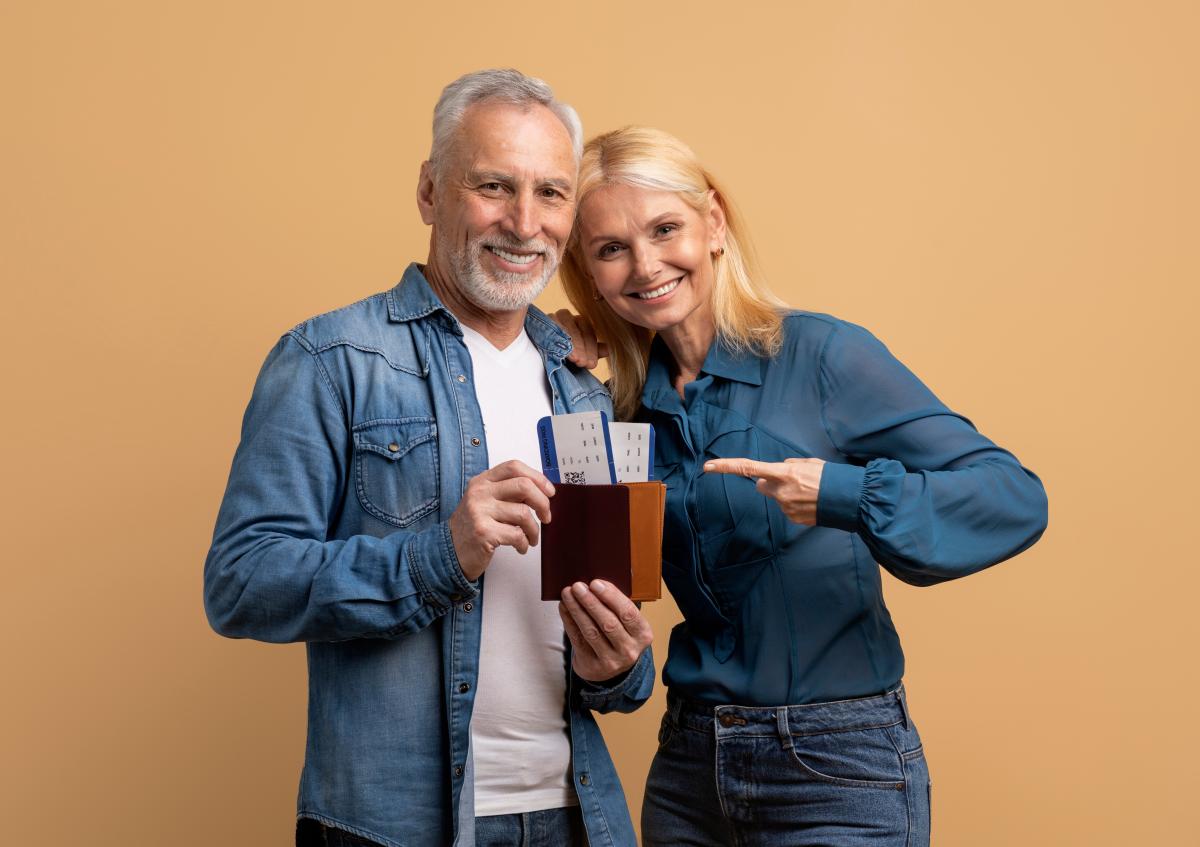 A smiling older man is holding a wallet with boarding passes inside, while a woman next to him is pointing at the wallet with an excited expression.
