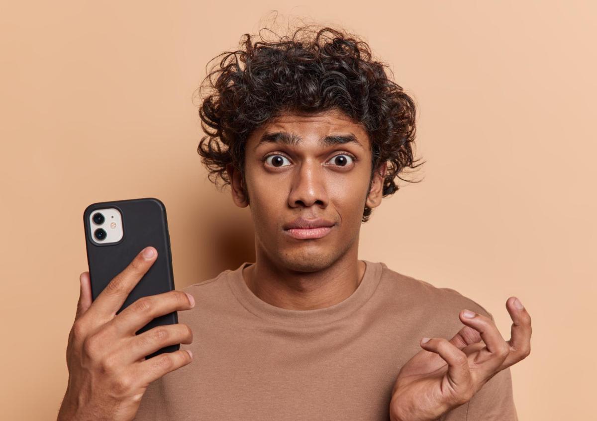 A man with a surprised and slightly concerned expression looks straight ahead while holding a mobile phone in one hand and raising his other hand with fingers slightly spread, as if he doesn’t understand something or is questioning it.