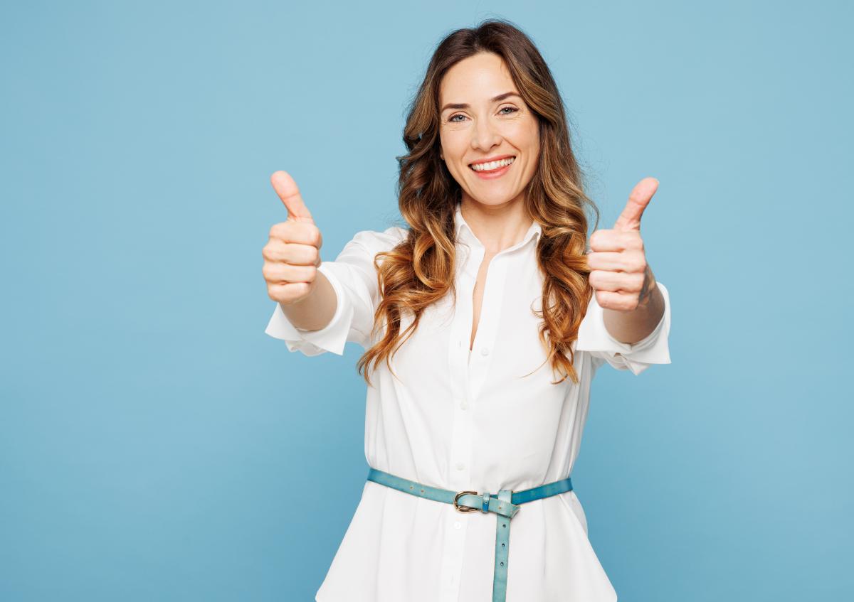 A smiling woman holding a laptop close to her chest while giving two thumbs up, expressing positivity and approval.
