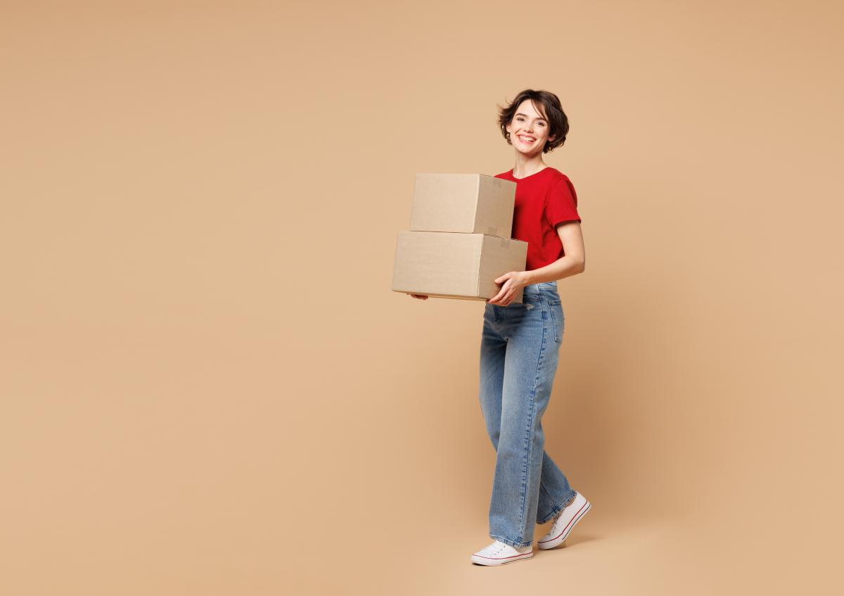 A woman smiling while carrying two stacked cardboard boxes, standing and looking forward.